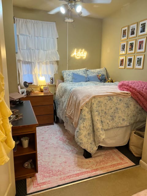 A cozy bedroom with a neatly made bed featuring floral bedding, a wall decorated with framed photographs, and a neon sign that reads "Addy" above the bed. There's a desk with a lamp, books, and decor, and two area rugs on the floor.